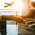 Traveler relaxes at airport while waiting for flight boarding, with plane taking off in background.