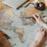 Hands marking destinations on a world map surrounded by travel essentials like passport and camera.