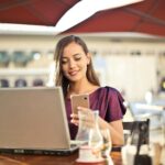 Woman enjoying remote work at a café, using a laptop and smartphone.