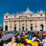 A large crowd gathers outside St. Peter's Basilica under a clear blue sky.