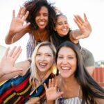 A group of diverse friends enjoying a sunny rooftop hangout, waving and smiling joyfully.